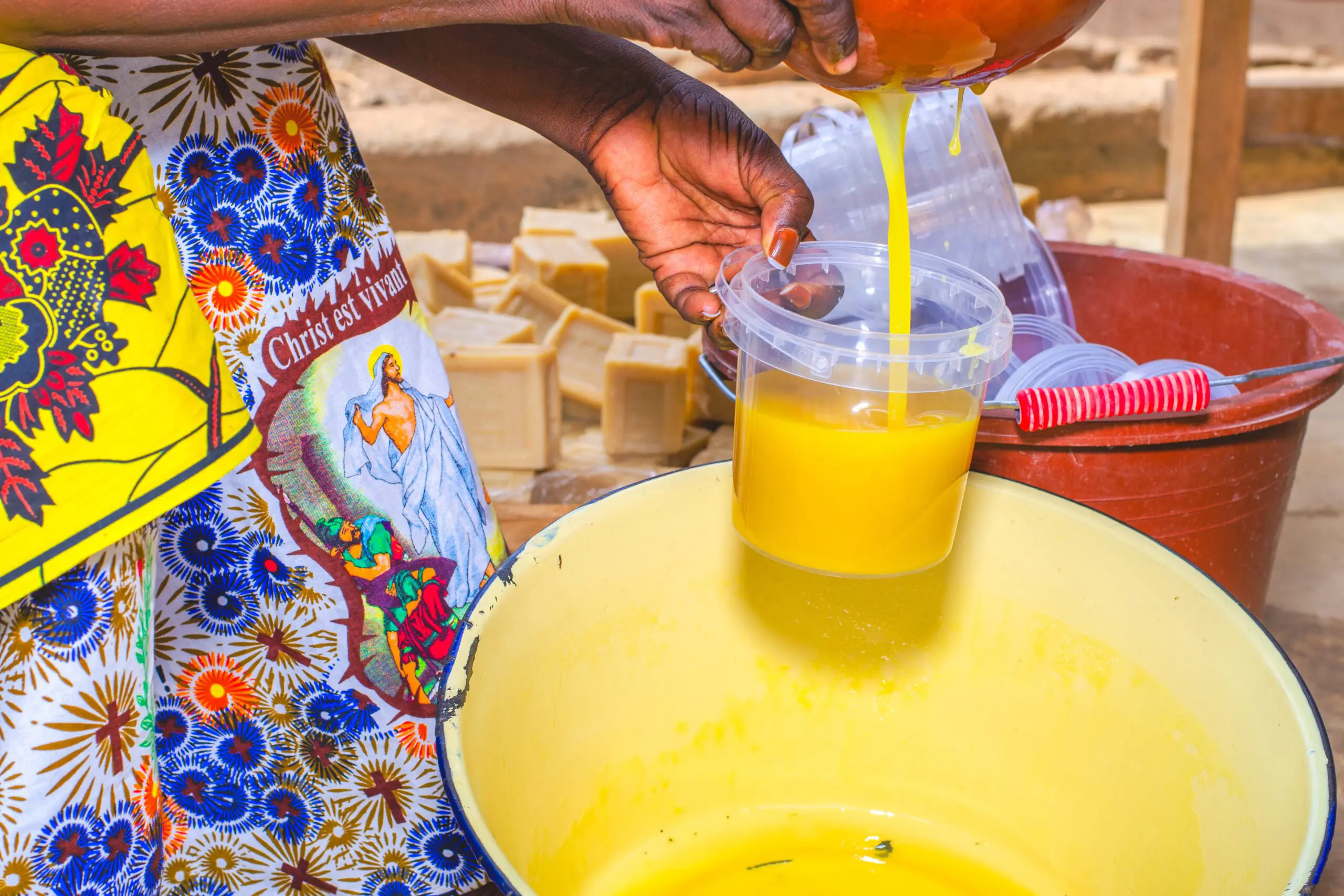 Eine Frau giesst leuchtend gelbe, flüssige Sheabutter aus einer Schüssel in einen transparenten Kunststoffbehälter. Im Hintergrund sind weitere leere Becher und rechteckige Seifenstücke zu sehen. Sie trägt ein farbenfrohes Kleid mit christlichem Motiv. Die Szene zeigt einen letzten Schritt in der Verarbeitungskette: die Abfüllung des fertigen Produkts für den Verkauf oder Gebrauch.