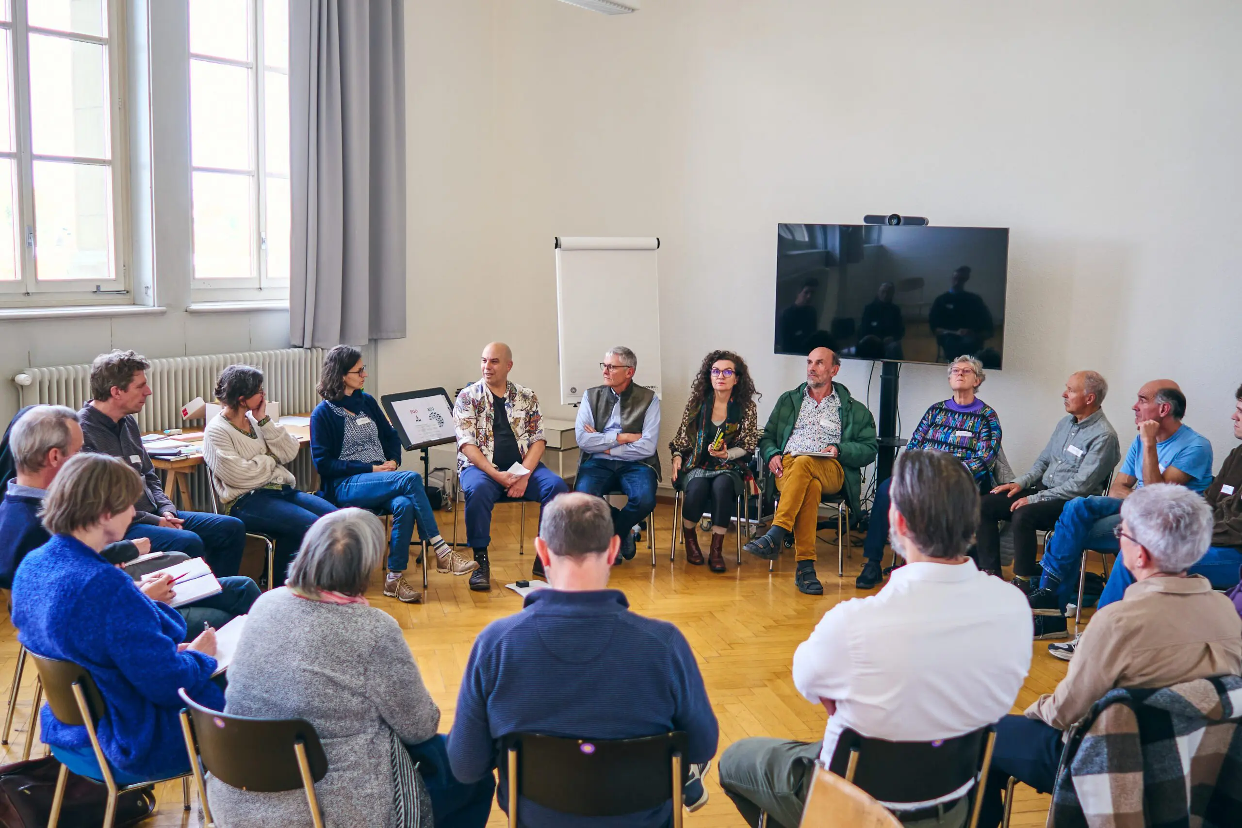 In einem hellen Seminarraum sitzen rund 20 Personen in einem großen Stuhlkreis. Die Gruppe ist bunt durchmischt: Männer und Frauen unterschiedlichen Alters, viele mit grauen Haaren, hören einander aufmerksam zu oder bereiten sich auf einen Redebeitrag vor. Der Austausch findet in ruhiger, konzentrierter Atmosphäre statt. Im Hintergrund stehen ein grosses schwarzes Display, ein Flipchart und ein aufgestelltes Poster. Der Raum ist schlicht gehalten mit Parkettboden, weißen Wänden und grossen Fenstern, durch die Tageslicht einfällt.