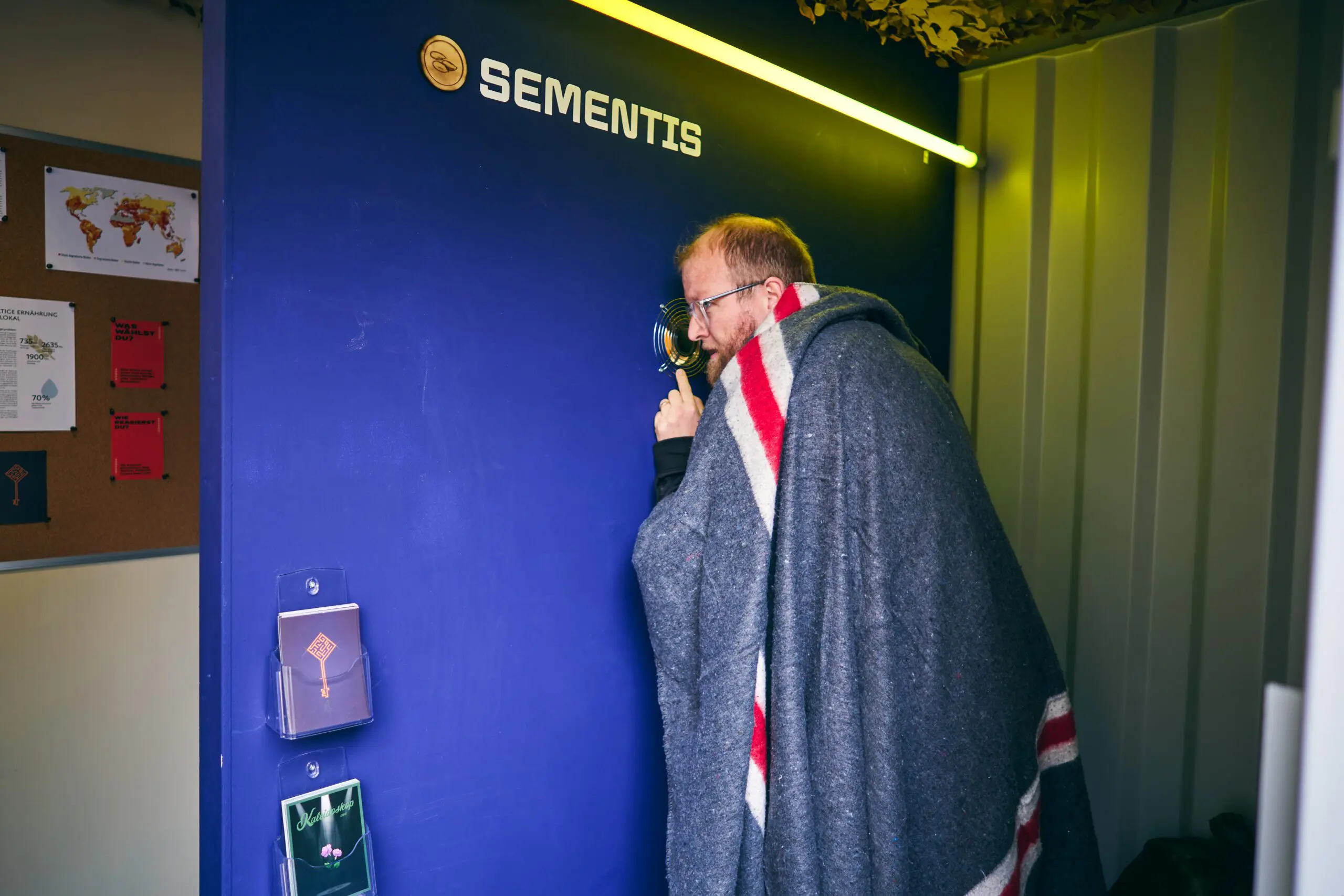 Ein Mann mit Brille und rotem Bart, eingehüllt in eine graue Decke mit roten und weißen Streifen, schaut durch ein Guckloch in eine blaue Wand mit der Aufschrift „Sementis“. Die Szene spielt sich in einem Escape Room Container ab. Im Hintergrund hängen Infotafeln mit globalen Statistiken und Weltkarten zur Ernährungssicherheit. Eine neonartige Lichtleiste beleuchtet die Wand von oben.