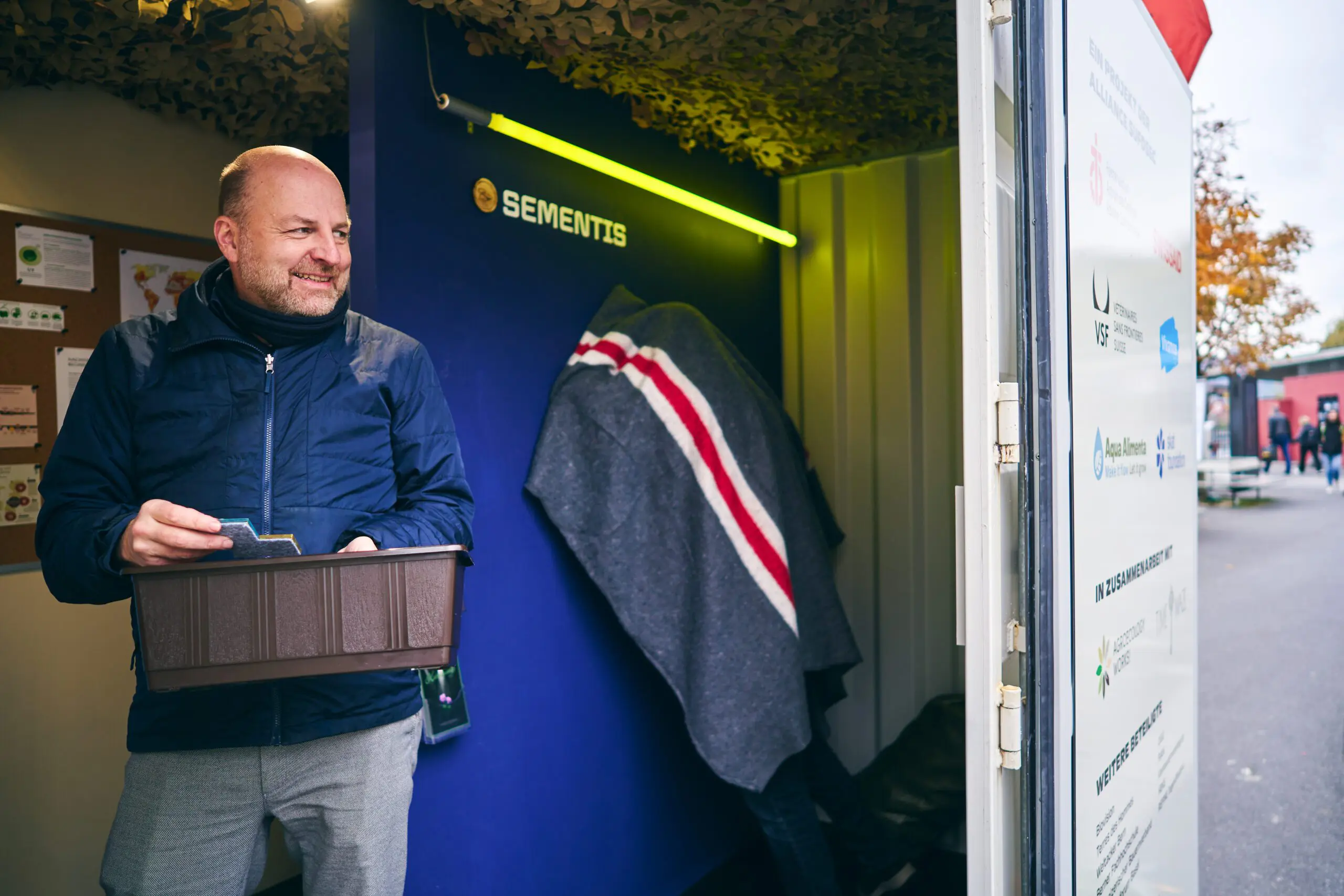 Ein Mann mit Glatze und blauer Jacke steht in einem Container vor einer blauen Wand mit der Aufschrift „Sementis“. Er hält einen braunen Pflanzkasten mit farbigen Holzplättchen darin und lächelt nach links. Hinter ihm kniet eine weitere Person mit einer grauen Decke mit roten und weißen Streifen über dem Rücken vor einem Guckloch in der Wand. Auf der rechten Seite ist ein Teil der Containertür mit Logos verschiedener Organisationen sichtbar.