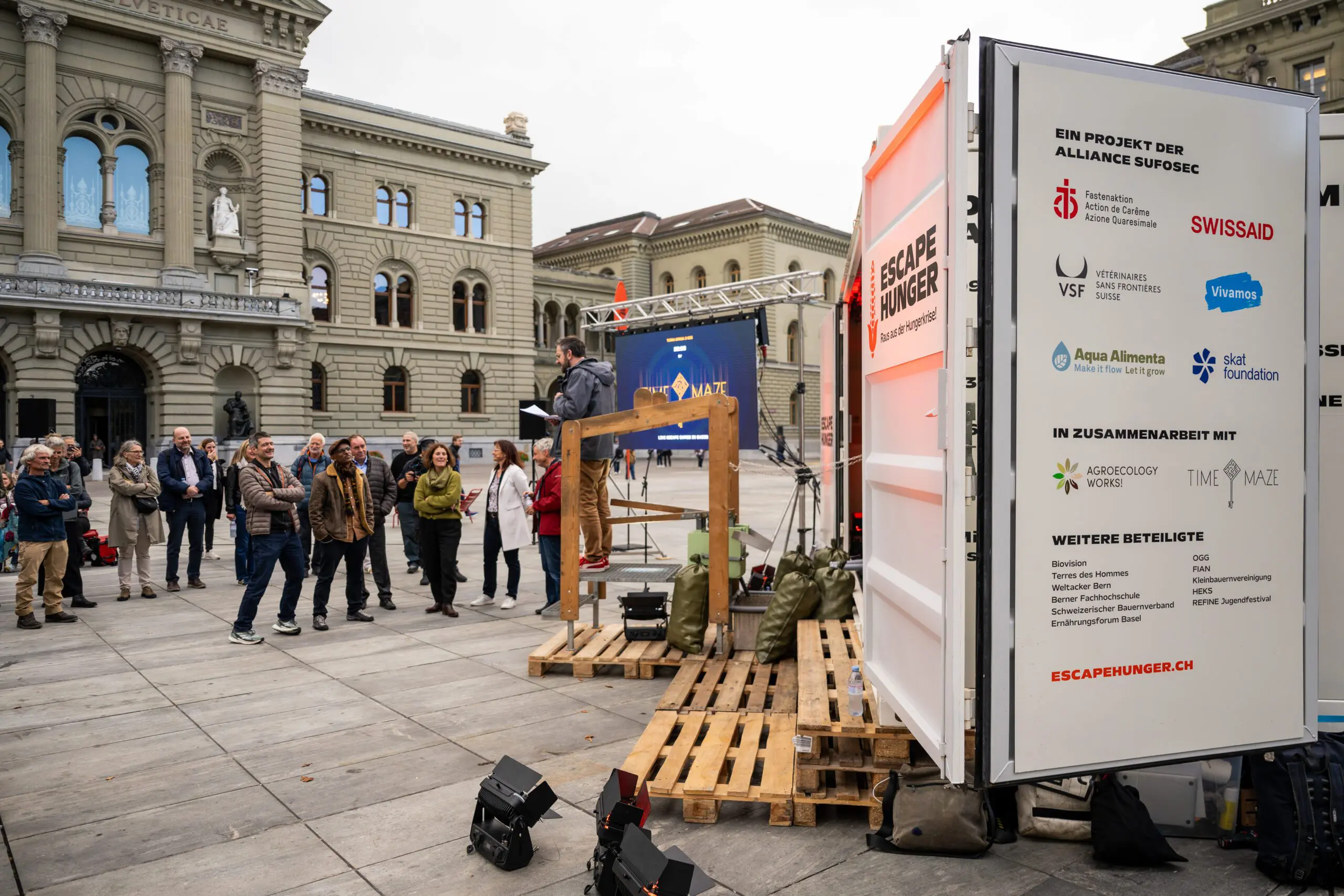 Eine Gruppe von Menschen steht auf dem Bundesplatz in Bern vor dem Bundeshaus. Im Vordergrund ist der „Escape Hunger“-Container geöffnet, daneben spricht ein Mann zu der Gruppe. Auf der Containerwand sind Logos von Fastenaktion, SWISSAID, VSF Suisse, Vivamos, Aqua Alimenta, Skat Foundation, Agroecology Works!, Time Maze und weiteren Partnerorganisationen sichtbar. Der Hintergrund zeigt die monumentale Fassade des Bundeshauses mit Säulen, Fenstern und der Inschrift „Curia Confederationis Helveticae“. Der Himmel ist bewölkt.