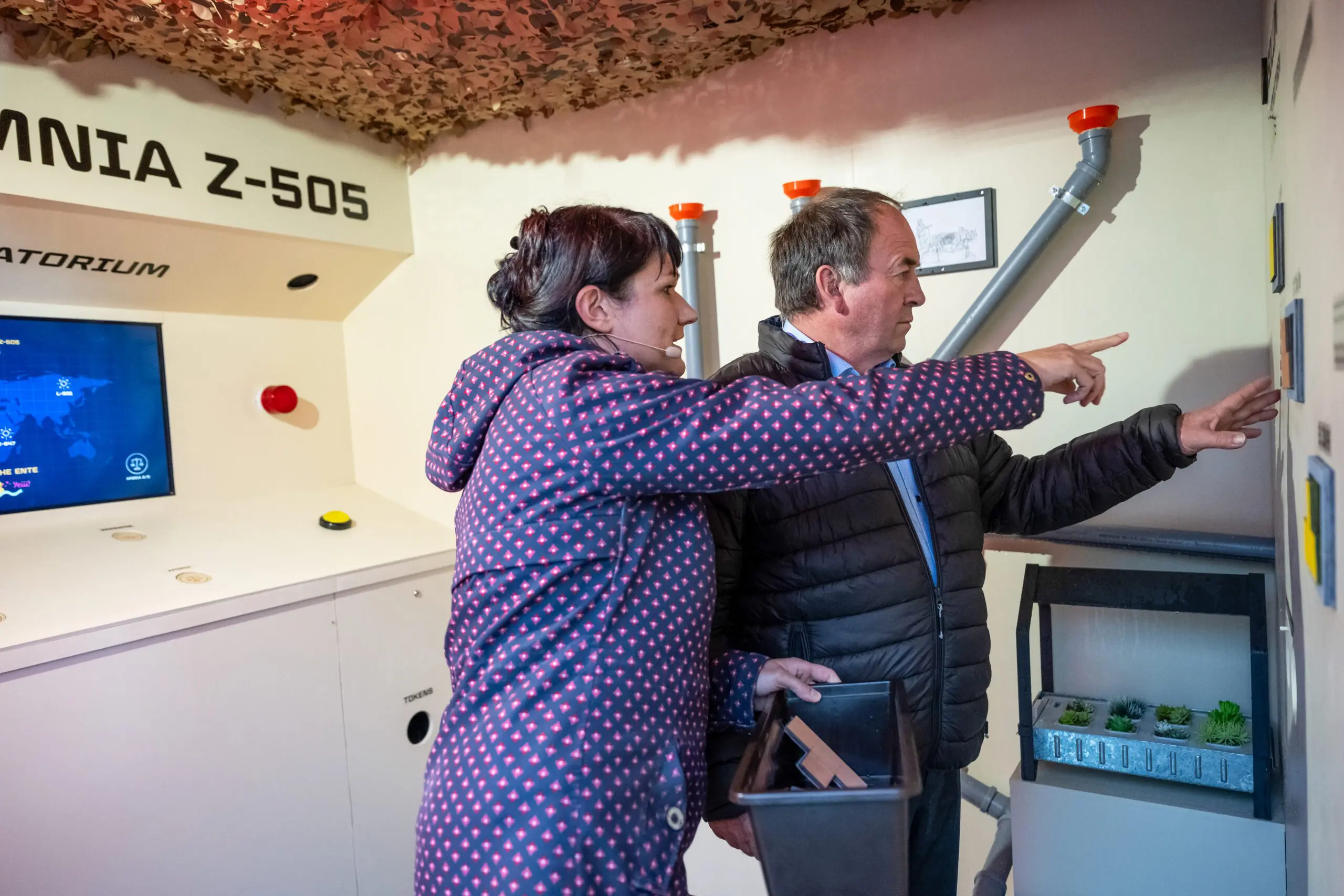Zwei Personen in warmer Kleidung stehen in einem Escape-Raum-Modul des Projekts „Escape Hunger“ und interagieren mit einer Wandinstallation. Sie bewegen kleine bunte Bausteine an eine beschriftete Tafel, die verschiedene landwirtschaftliche Szenarien darstellt. Im Hintergrund sind Rohre und Informationsposter zur globalen Ernährungssituation sichtbar. Die Szene vermittelt eine interaktive Lernerfahrung zu nachhaltiger Landwirtschaft in einem kreativ gestalteten Raum mit Tarnnetzdecke.