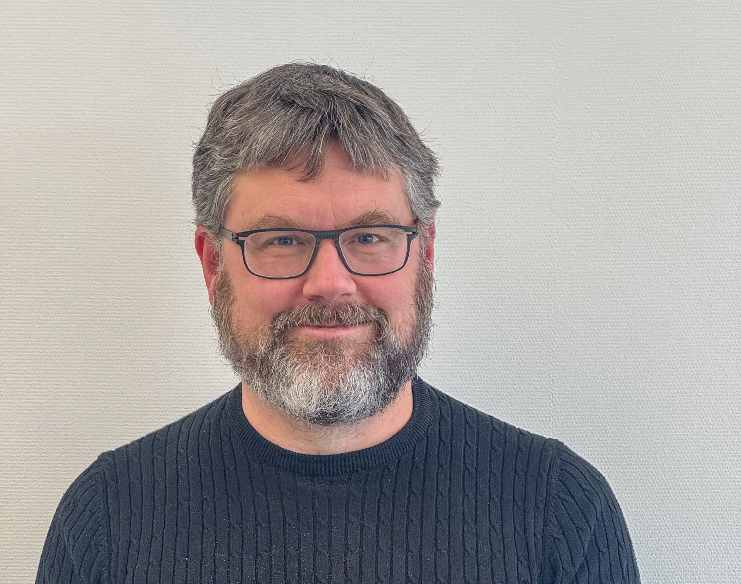 Ein Mann mit graumeliertem Haar und Bart, Brille und dunkelblauem Pullover mit feinem Strickmuster. Er steht vor einer hellen Wand und lächelt freundlich in die Kamera.
