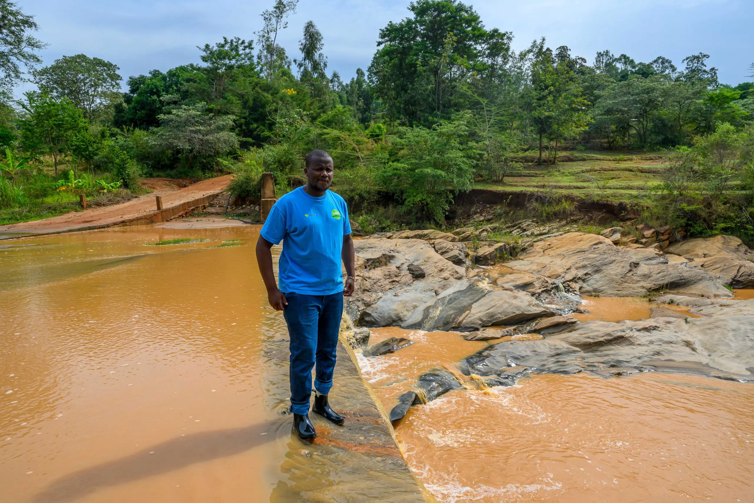 Ein Mann steht in Gummistiefeln auf einer niedrigen Staustufe in einem Fluss mit braunem Wasser. Umgeben von Felsen, Bäumen und landwirtschaftlich genutzten Flächen betrachtet er die Wasserführung. Im Hintergrund führt eine kleine Brücke über den Fluss. Die Szene verdeutlicht die Bedeutung von Wasserressourcen und Bewässerung für die Landwirtschaft in einer ländlichen Region.