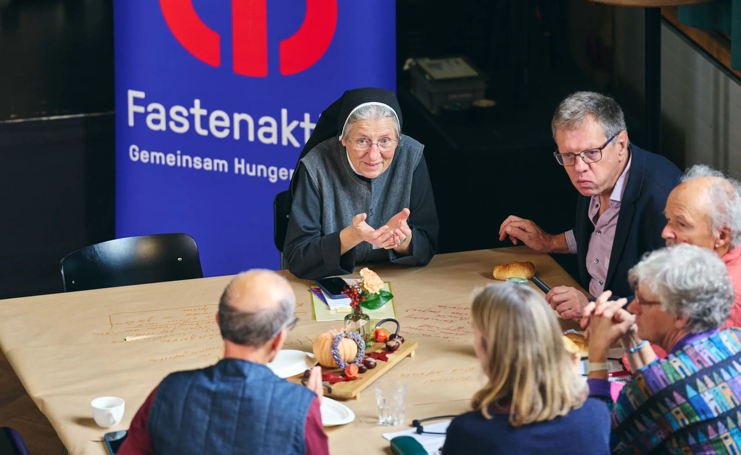 Eine Ordensschwester mit Brille und schwarzem Habit diskutiert am Aktionsforum von Fastenaktion mit fünf weiteren Teilnehmenden an einem Tisch. Sie gestikuliert mit beiden Händen, während die anderen aufmerksam zuhören. Auf dem mit Papier bedeckten Tisch stehen Getränke, kleine Speisen und eine herbstliche Dekoration. Im Hintergrund ist ein blaues Banner mit dem Logo von Fastenaktion zu sehen.