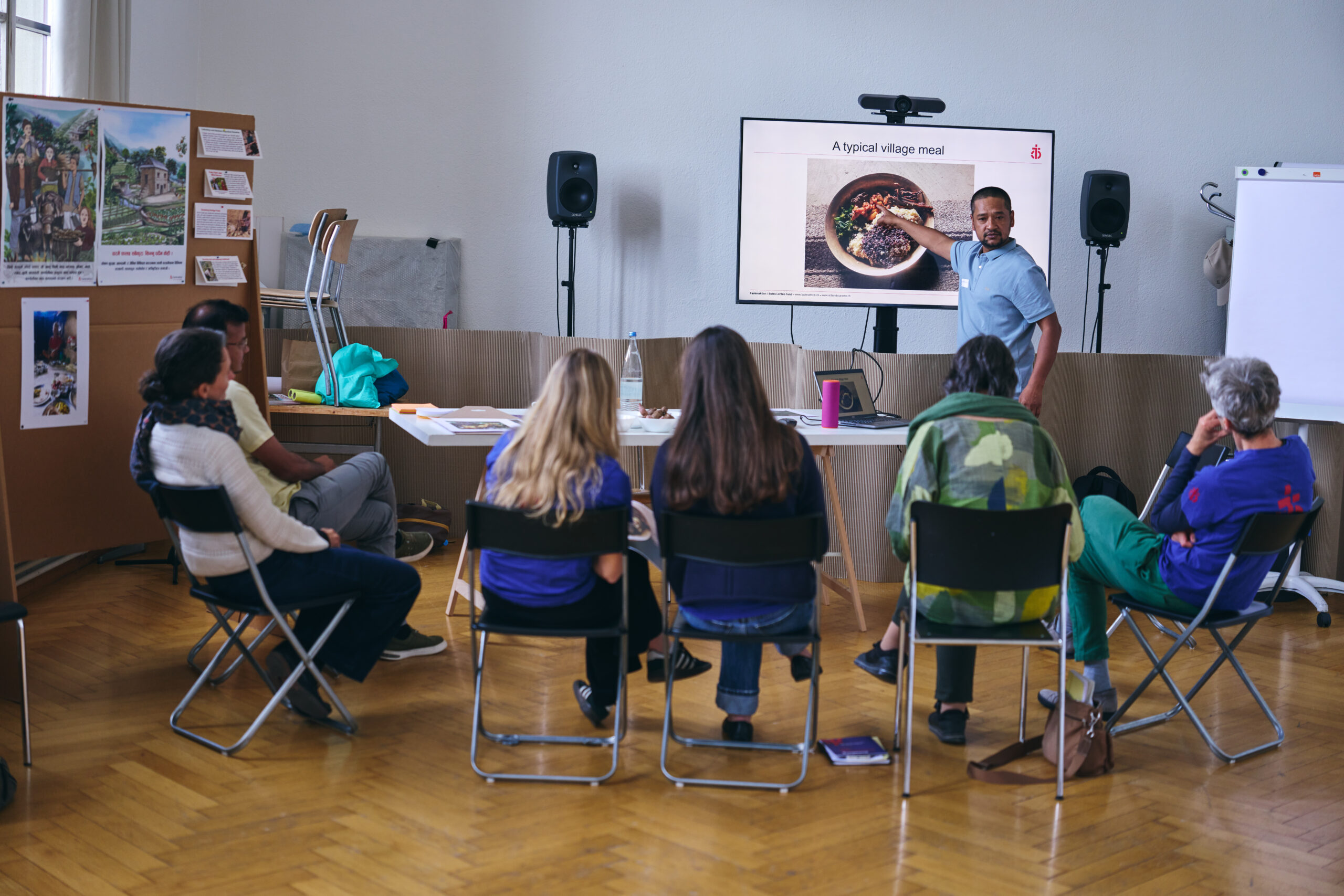 Ein Mann im hellblauen Poloshirt hält an einem Workshop des Aktionsforums von Fastenaktion eine Präsentation über eine typische Mahlzeit in einem Dorf. Er zeigt auf ein Foto mit einem Teller Essen auf dem Bildschirm. Sechs Teilnehmende sitzen im Halbkreis und hören zu. Links stehen Stellwände mit Fotos und Informationen zum Leben in ländlichen Gemeinschaften.